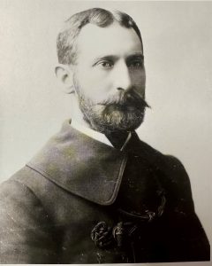 A portrait of Charles Woodruff, Fort Mackinac's Assistant Post Surgeon, in 1895. The photo is black and white, and he has a beard and large mustache, and is wearing an overcoat with a large collar. 