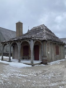 The Post Guardhouse at Colonial Michilimackinac. The building is gray, with a wood shingle roof, with pillars in front. The ground in front is gravel and dirt, with a light dusting of snow. 