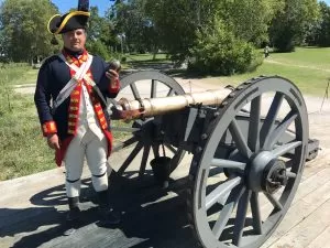 Light bronze 6-pound guns, mounted on highly mobile traveling carriages, made up the part of the early British defenses of Fort Mackinac. Here, a bombardier of the Royal Regiment of Artillery holds a 6-pound cannonball, which gives the weapon its name.